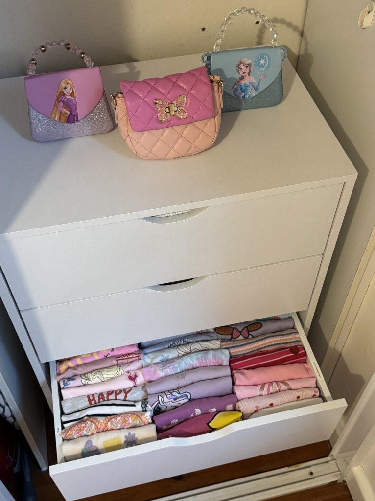 A neatly organized drawer filled with folded children's clothing in various shades of pink and purple, alongside three colorful toy bags on top of a white dresser.